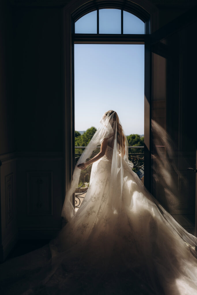 Bride in a lace gown and long veil standing at an arched window overlooking treetops