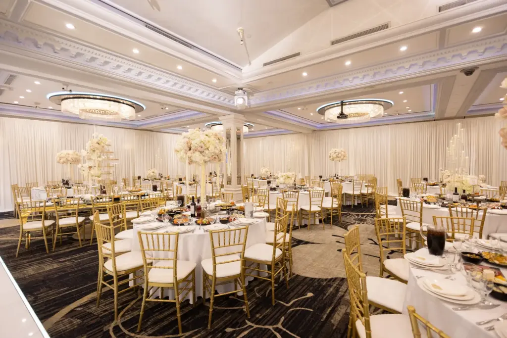 Wide view of ballroom reception with round tables, gold chiavari chairs and crystal chandeliers