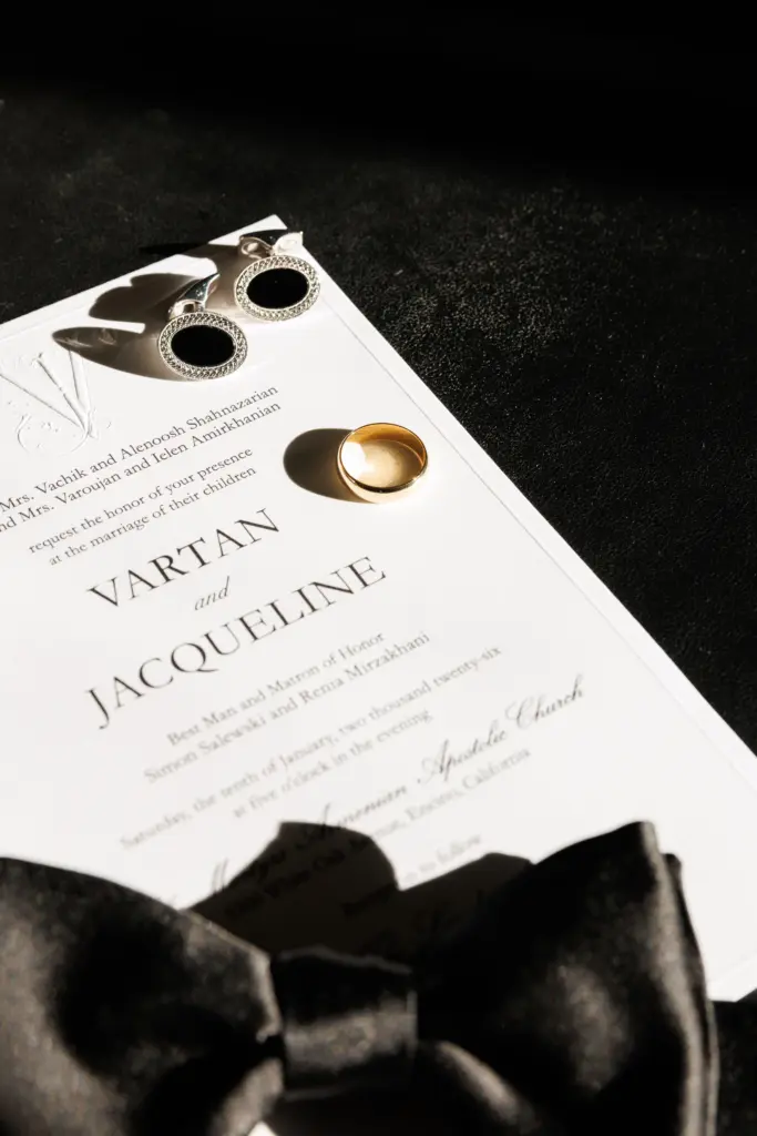 Wedding invitation with a gold ring, cufflinks and a black bow tie on a dark background