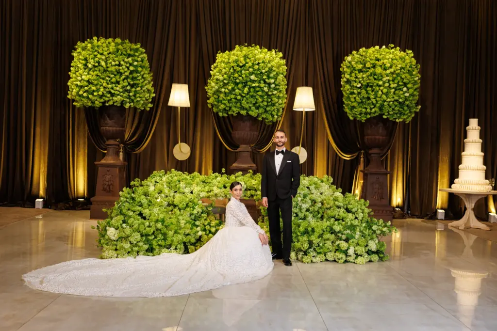 Bride in a lace gown and groom in a black tuxedo posing beside tall green topiary trees