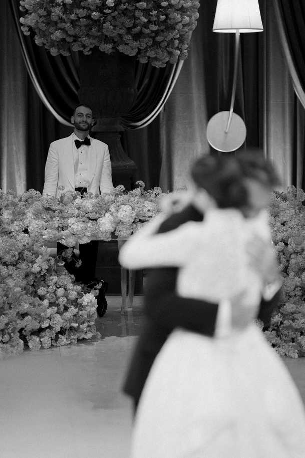Black-and-white photo of a groom watching a dancing couple behind a floral arrangement
