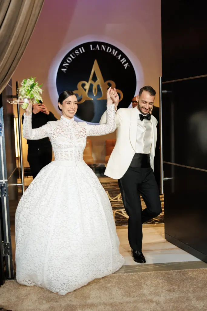 Bride and groom celebrating with raised hands beneath an illuminated venue logo at the entrance