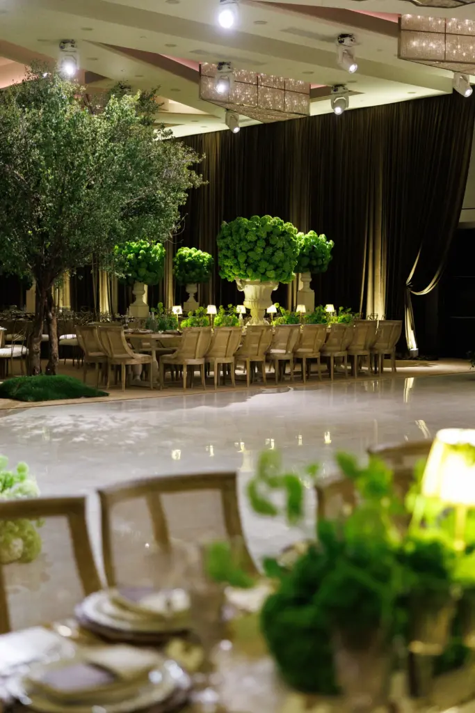 Long reception table with green floral garlands and wooden chairs beneath an indoor olive tree