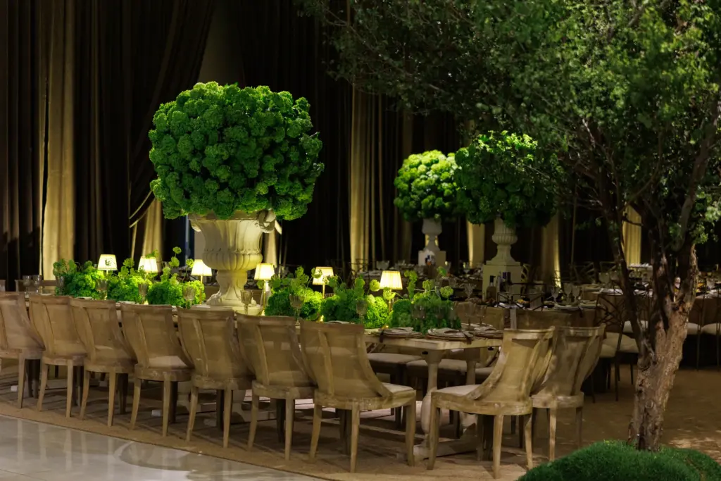 Long banquet table set for guests beside a tall topiary centerpiece and tiered cake