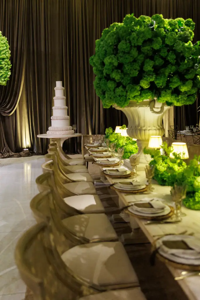 Bride and groom posing beside a floral hedge with a tiered wedding cake nearby