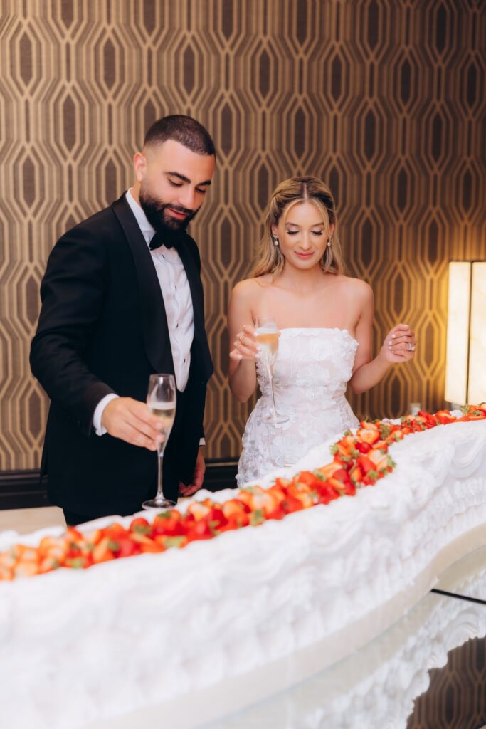 Bride and groom toasting with champagne beside their strawberry-topped wedding cake