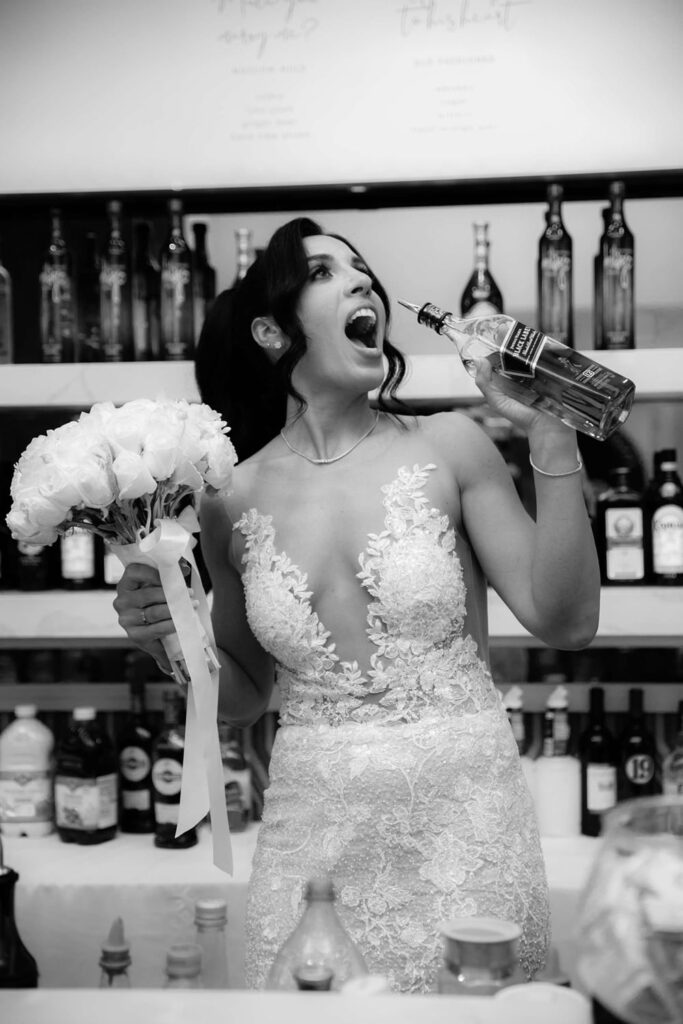 Black and white portrait of the bride taking a playful shot behind the bar
