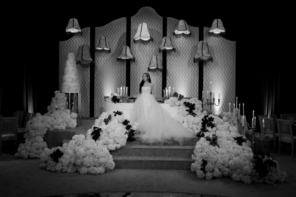 Black-and-white photo of a bride standing on a flower-lined stage in a ballroom