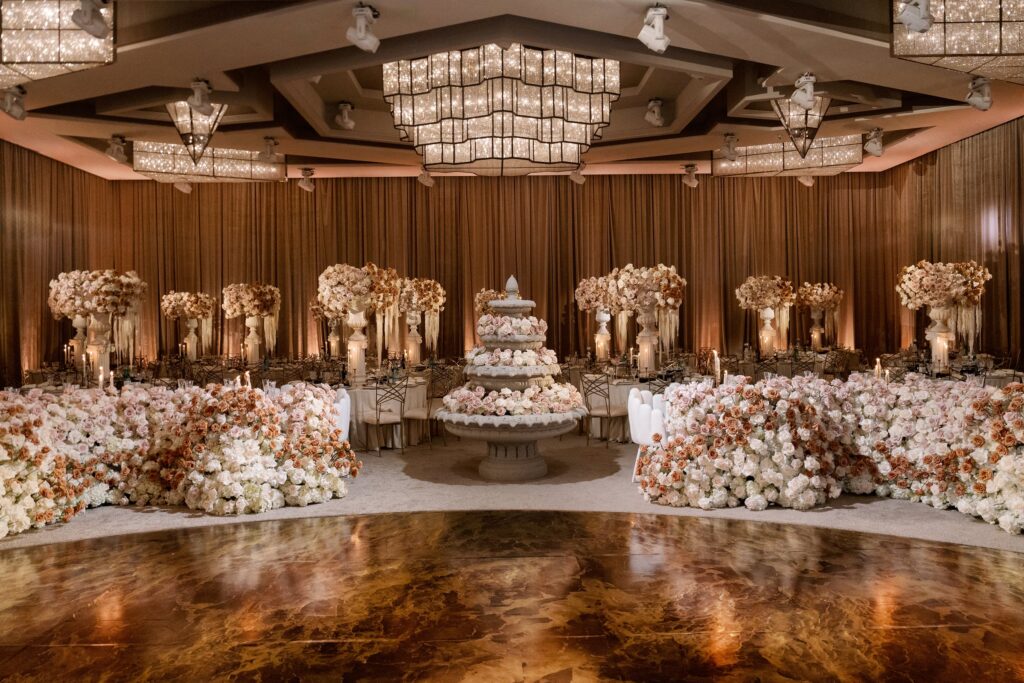 Wide view of a ballroom reception with pink-toned floral urns flanking a tiered wedding cake