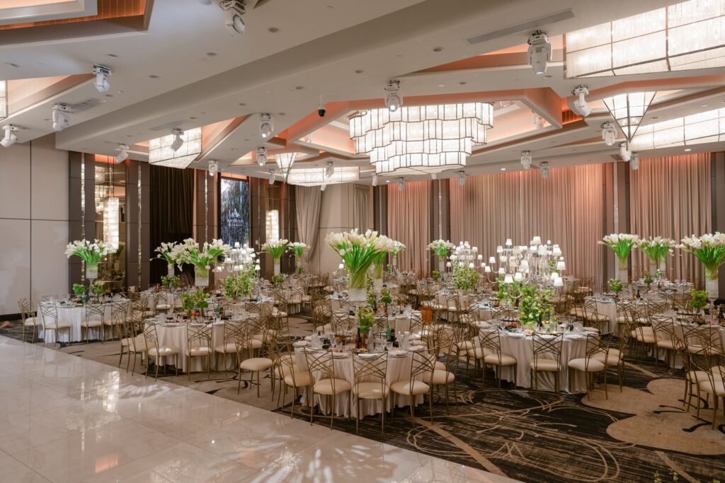 Banquet hall set for a reception with round tables, tall white lily centerpieces, and gold chairs