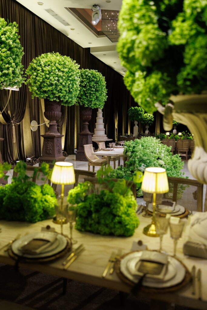 Reception table with a green floral centerpiece and lamp beside topiary trees and a tiered cake