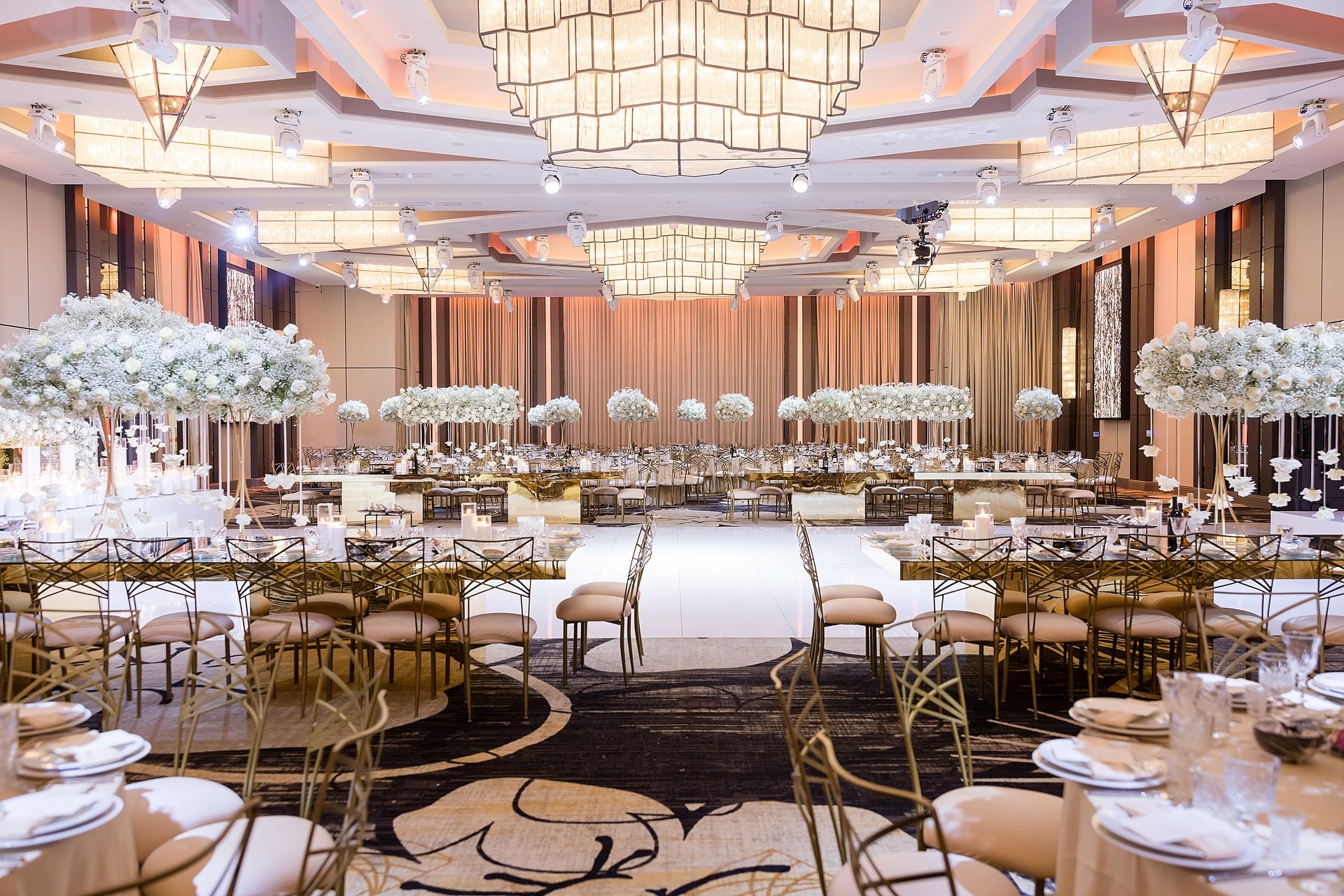 Ballroom reception with a white dance floor aisle framed by tall white floral arrangements
