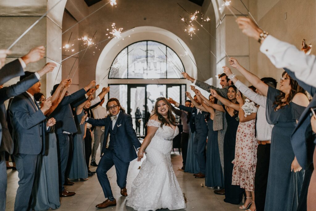 Bride and groom exit under crossed sparklers held by cheering guests at a stone archway