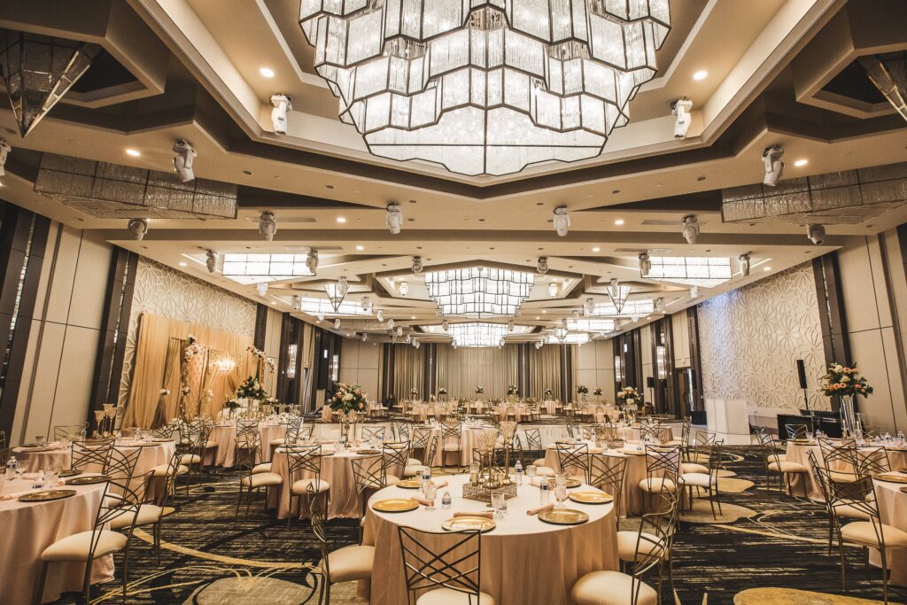 Banquet hall with blush-linen round tables, gold chairs, and a floral stage backdrop