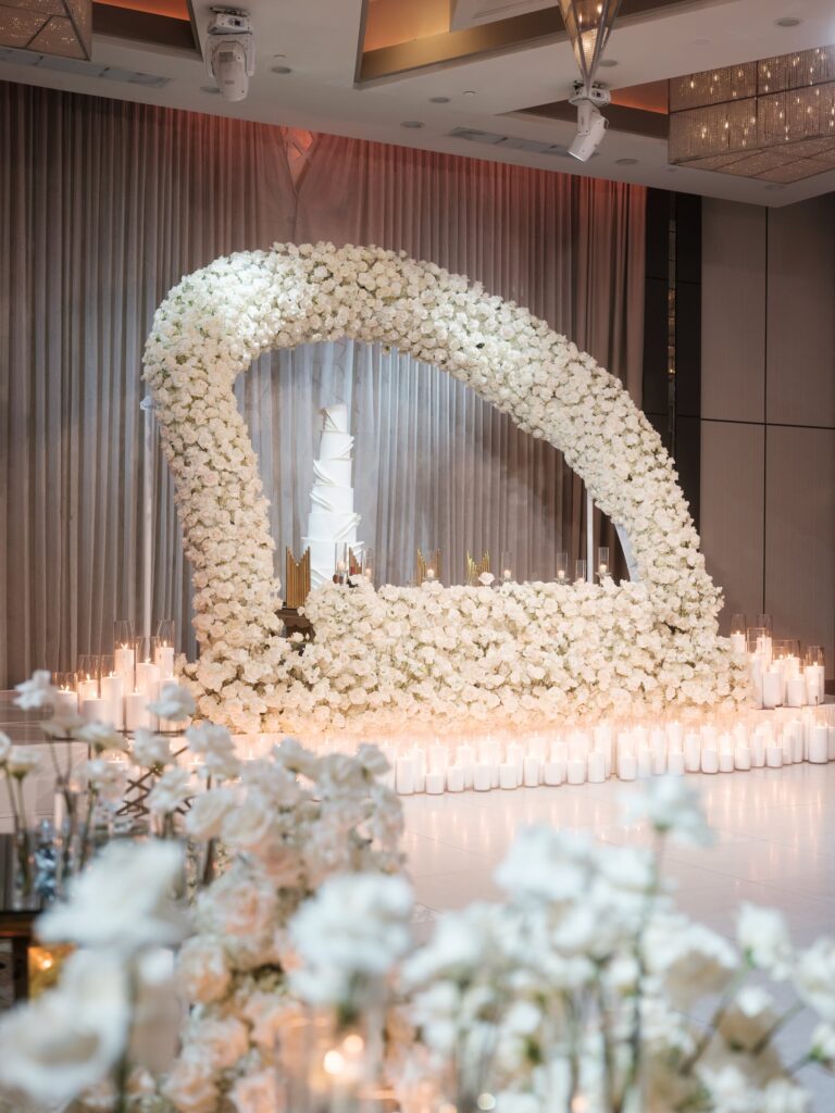 White rose arch framing a tiered wedding cake surrounded by rows of pillar candles