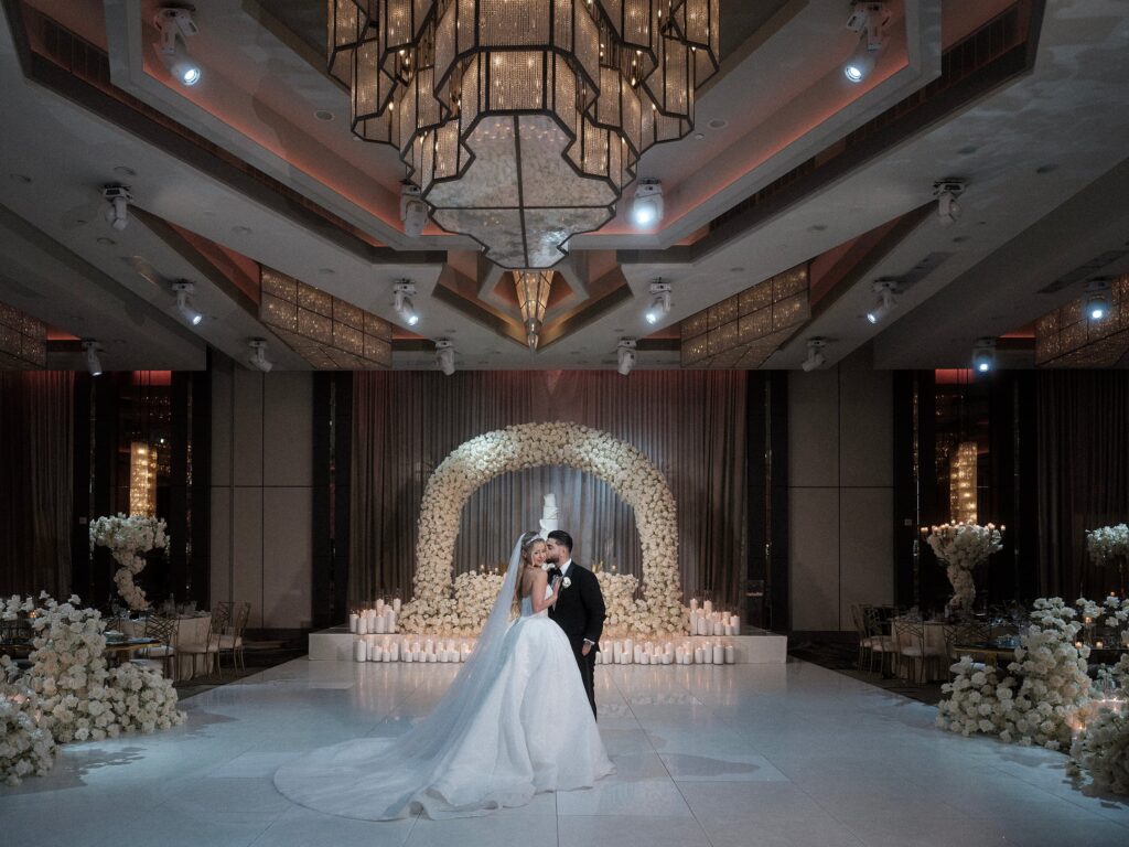 Bride and groom embrace under a floral arch on a candlelit dance floor at night