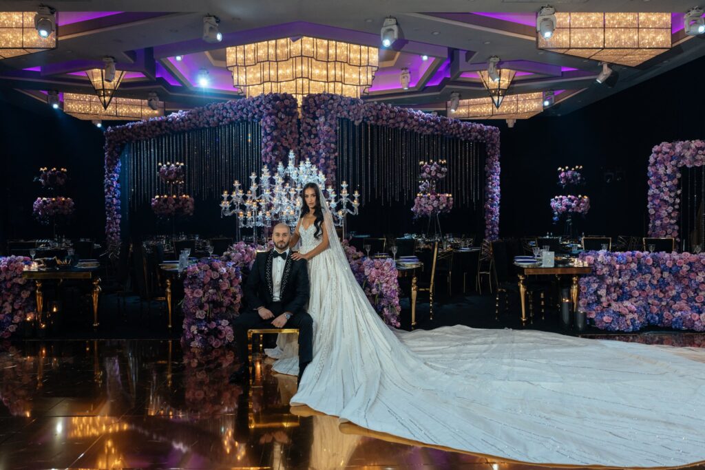 Bride and groom pose beside a gold bench in front of a purple floral backdrop and chandelier