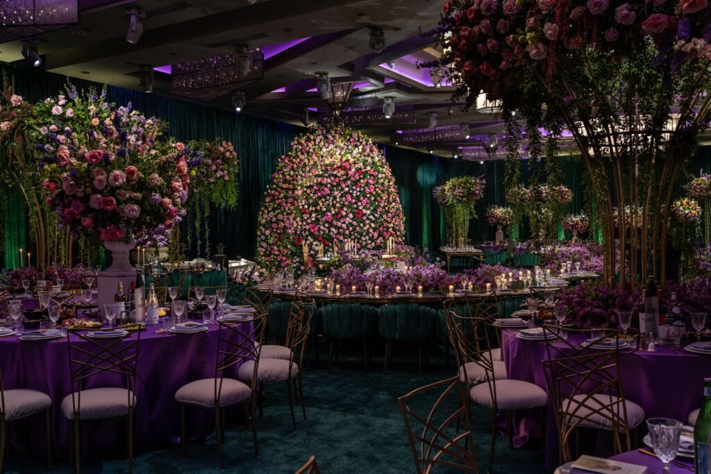 Purple-lit reception hall with a floral installation over the head table and teal chairs