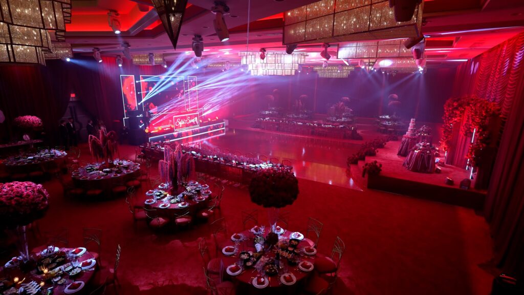 Red-lit reception hall with a stage sign and round tables ready for a celebration