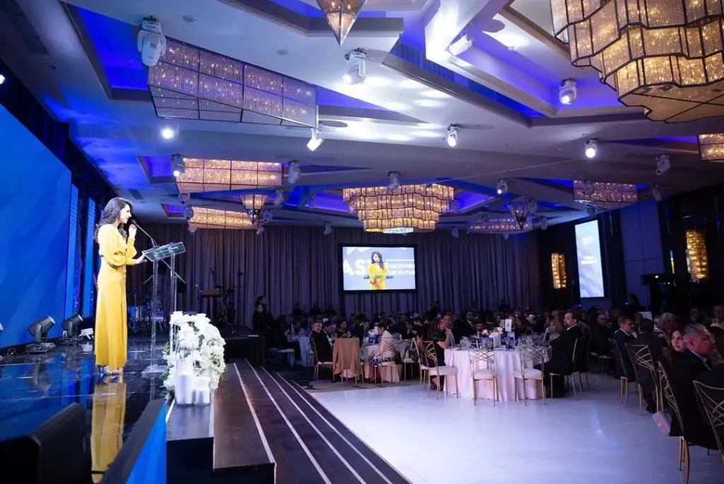 Woman speaking at a podium during a gala event with a large screen behind her