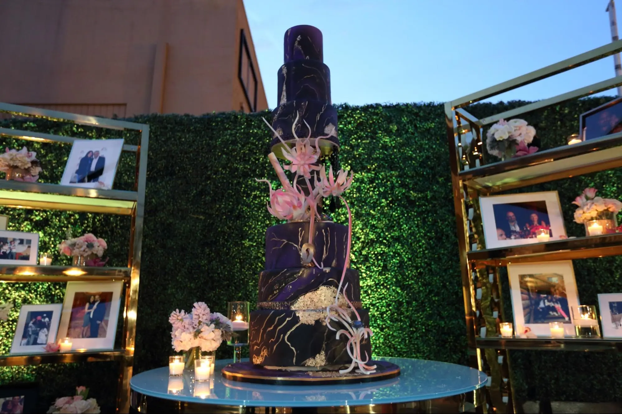 Tall purple wedding cake with pink floral accents displayed among photo shelves