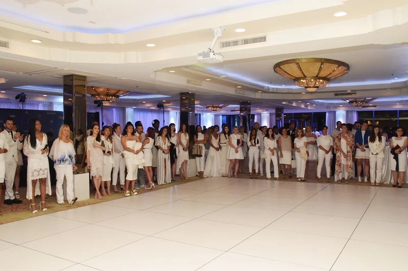 Guests dressed in white lined up along a ballroom aisle beneath crystal chandeliers