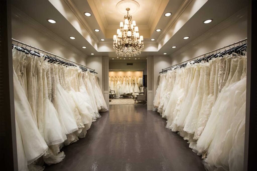 Aisle of white wedding gowns on display beneath a chandelier in a bridal boutique