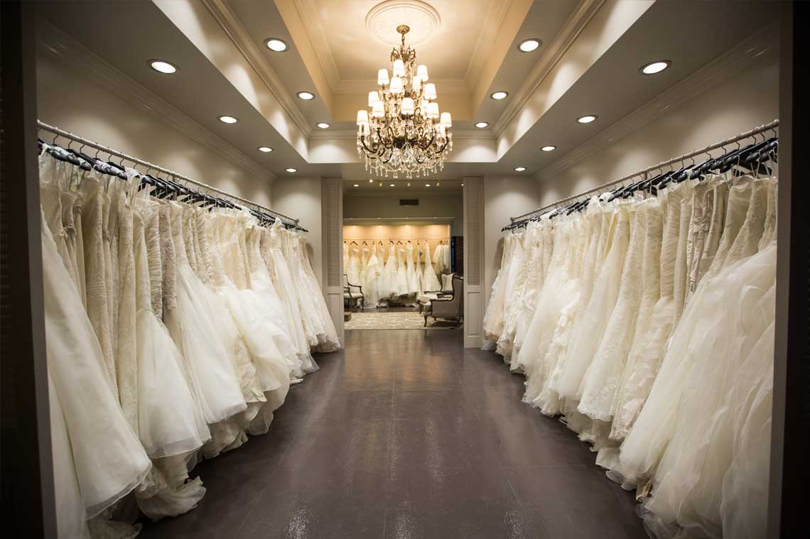 Aisle of white wedding gowns on display beneath a chandelier in a bridal boutique