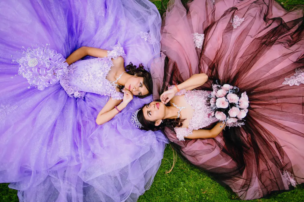 Two girls in purple and pink quinceañera gowns lying on grass with bouquets