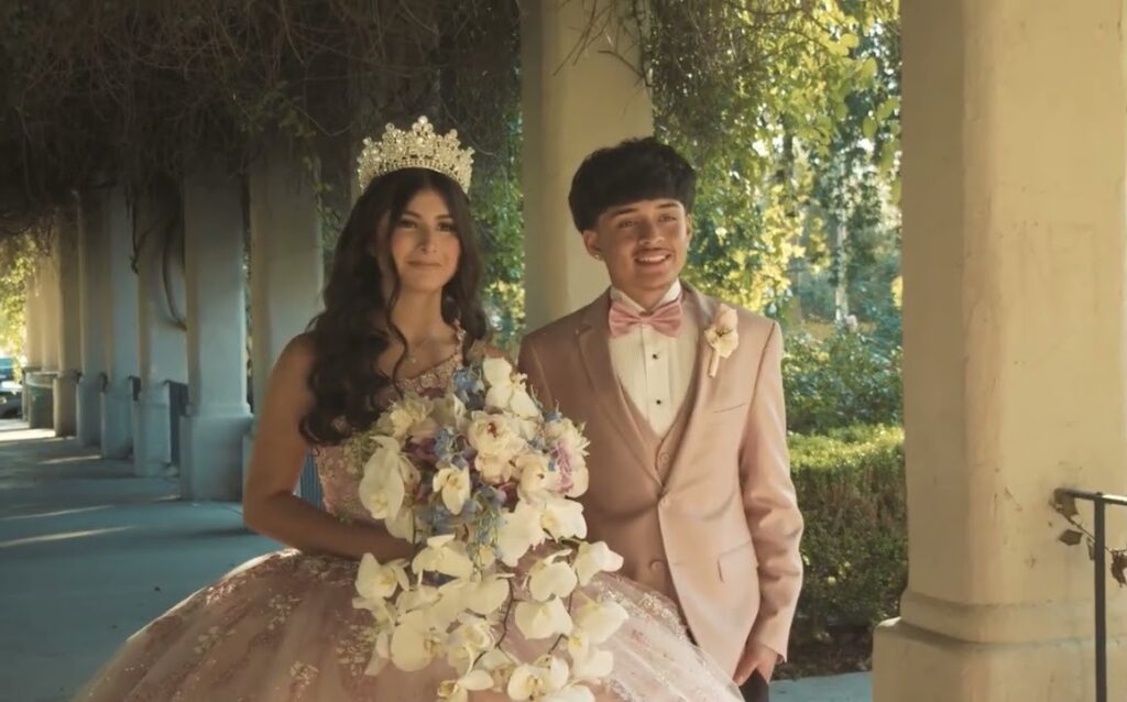 Quinceañera in a pink gown and tiara standing with an escort under a colonnade
