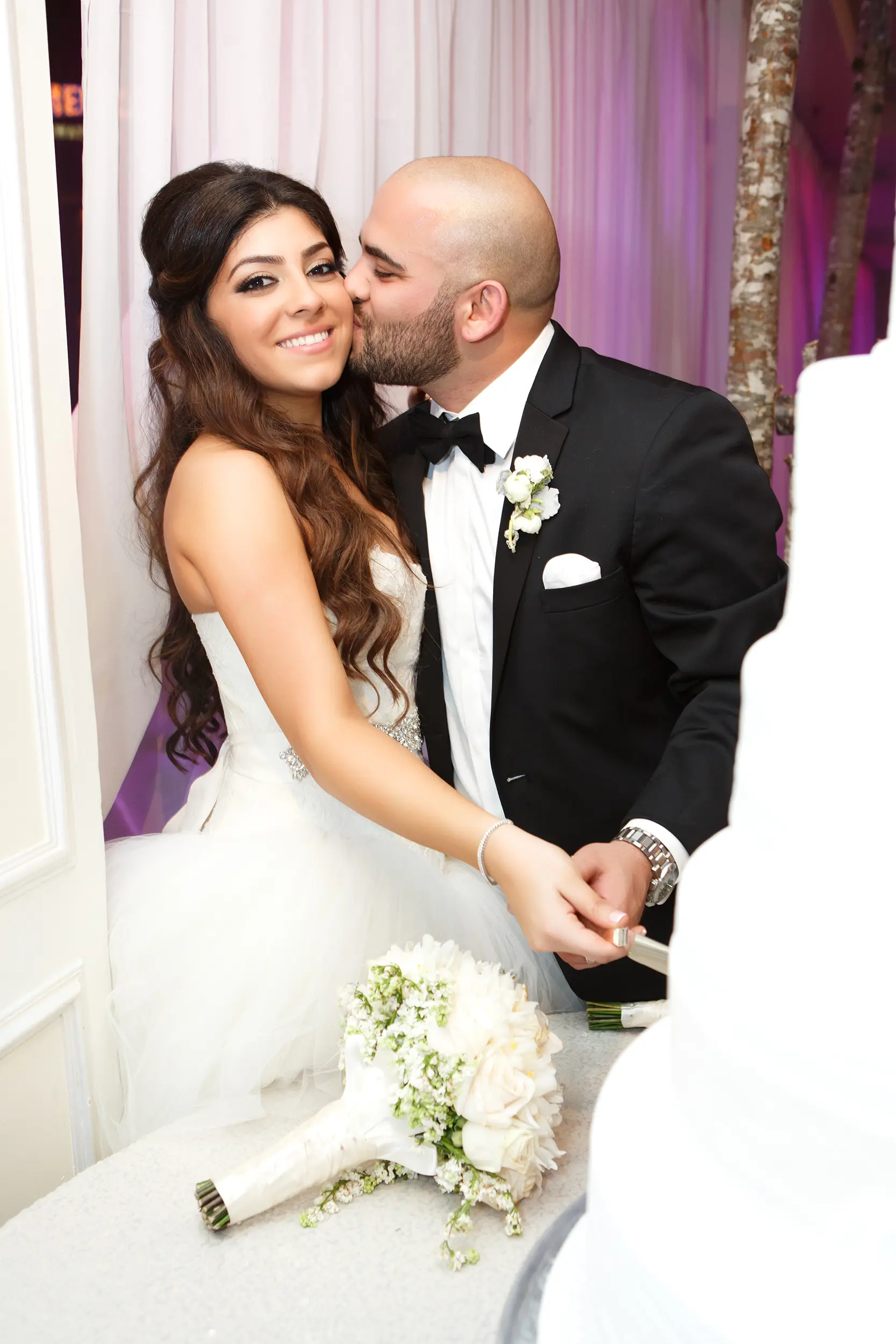 Groom kissing bride's cheek beside the wedding cake at their reception