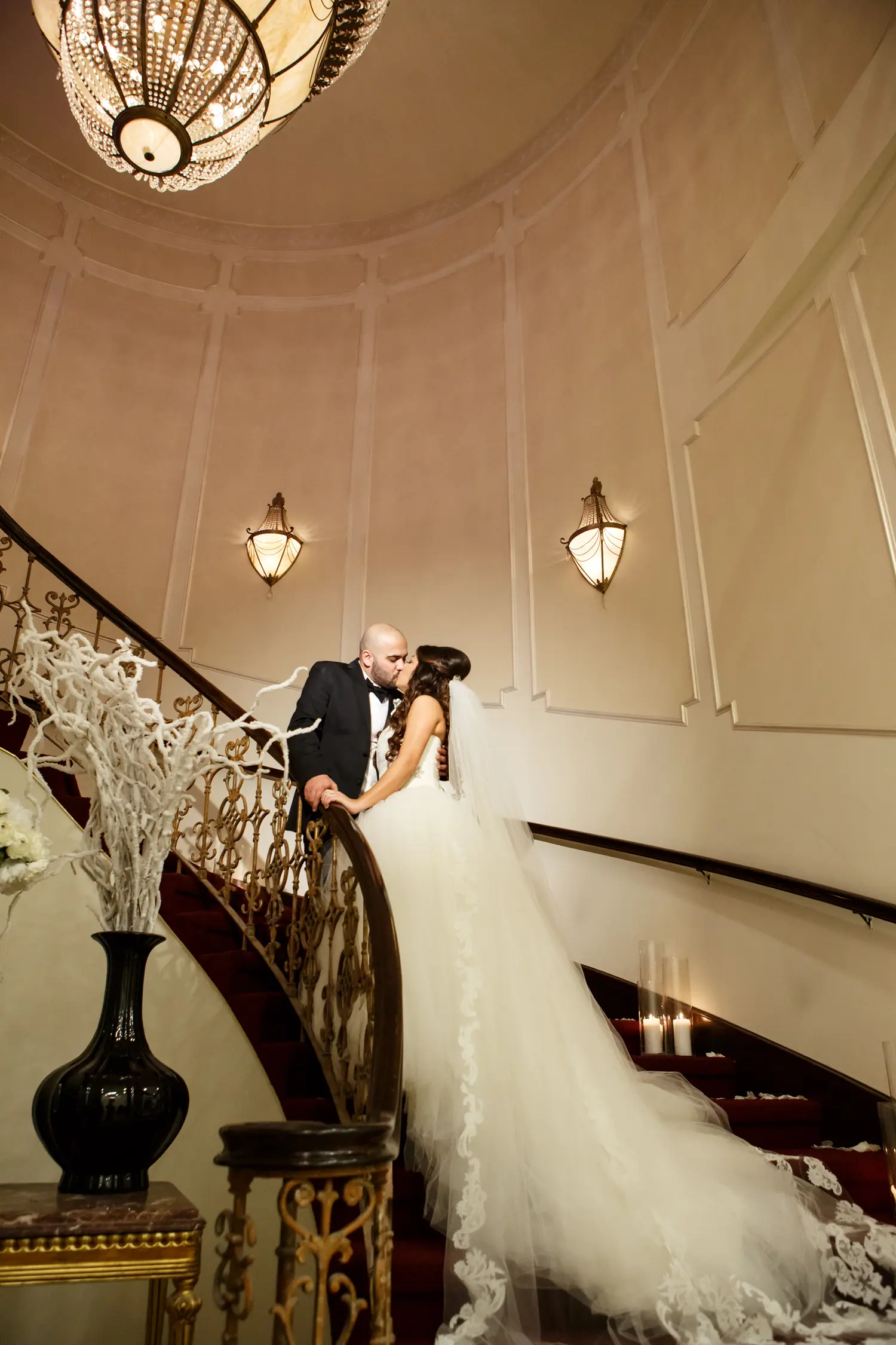 Bride and groom kissing on a grand staircase beneath a chandelier