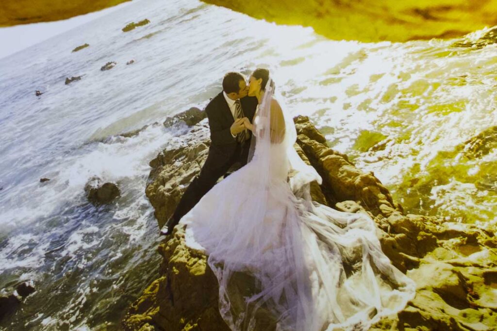 Bride and groom kissing on ocean-side rocks at sunset