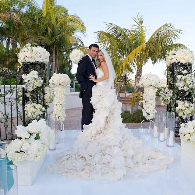 Bride in a ruffled gown and groom embracing at an outdoor floral ceremony arch with palm trees