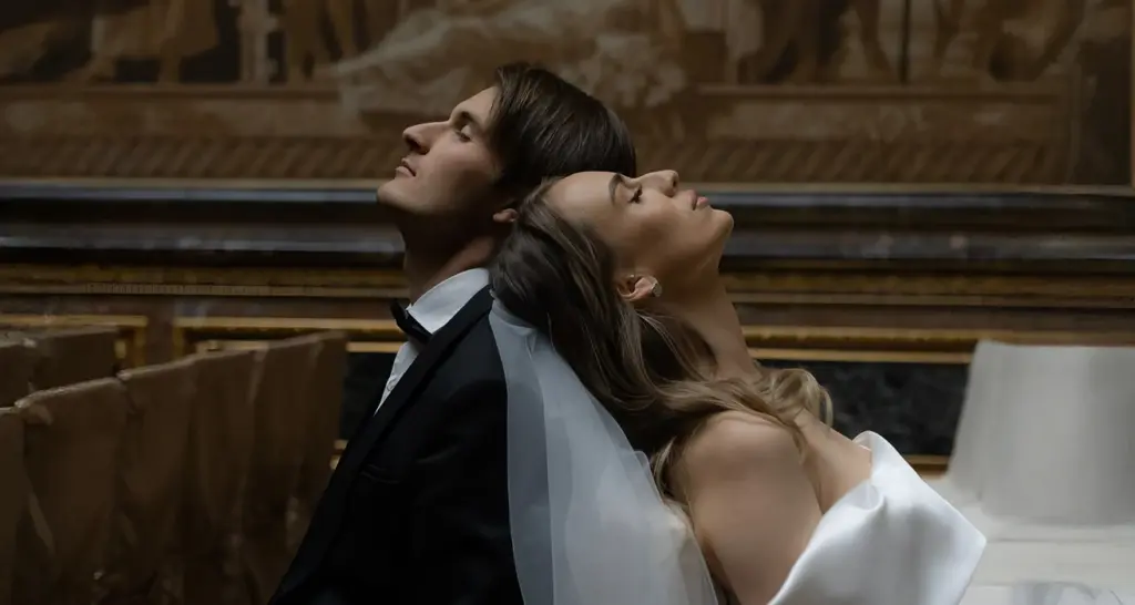 Bride and groom lean back together in a dramatic pose inside a dim chapel