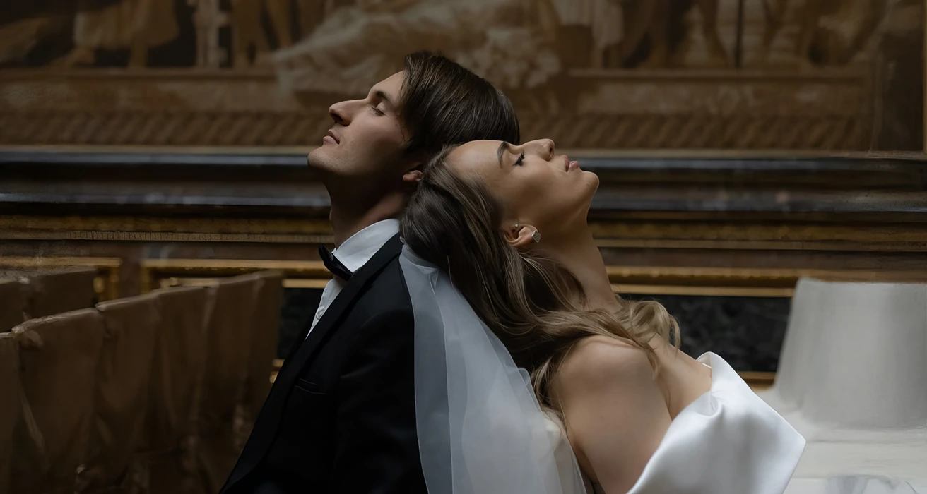 Bride and groom lean back together in a dramatic pose inside a dim chapel