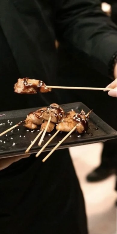 Server presenting grilled chicken skewers with teriyaki glaze on a black plate
