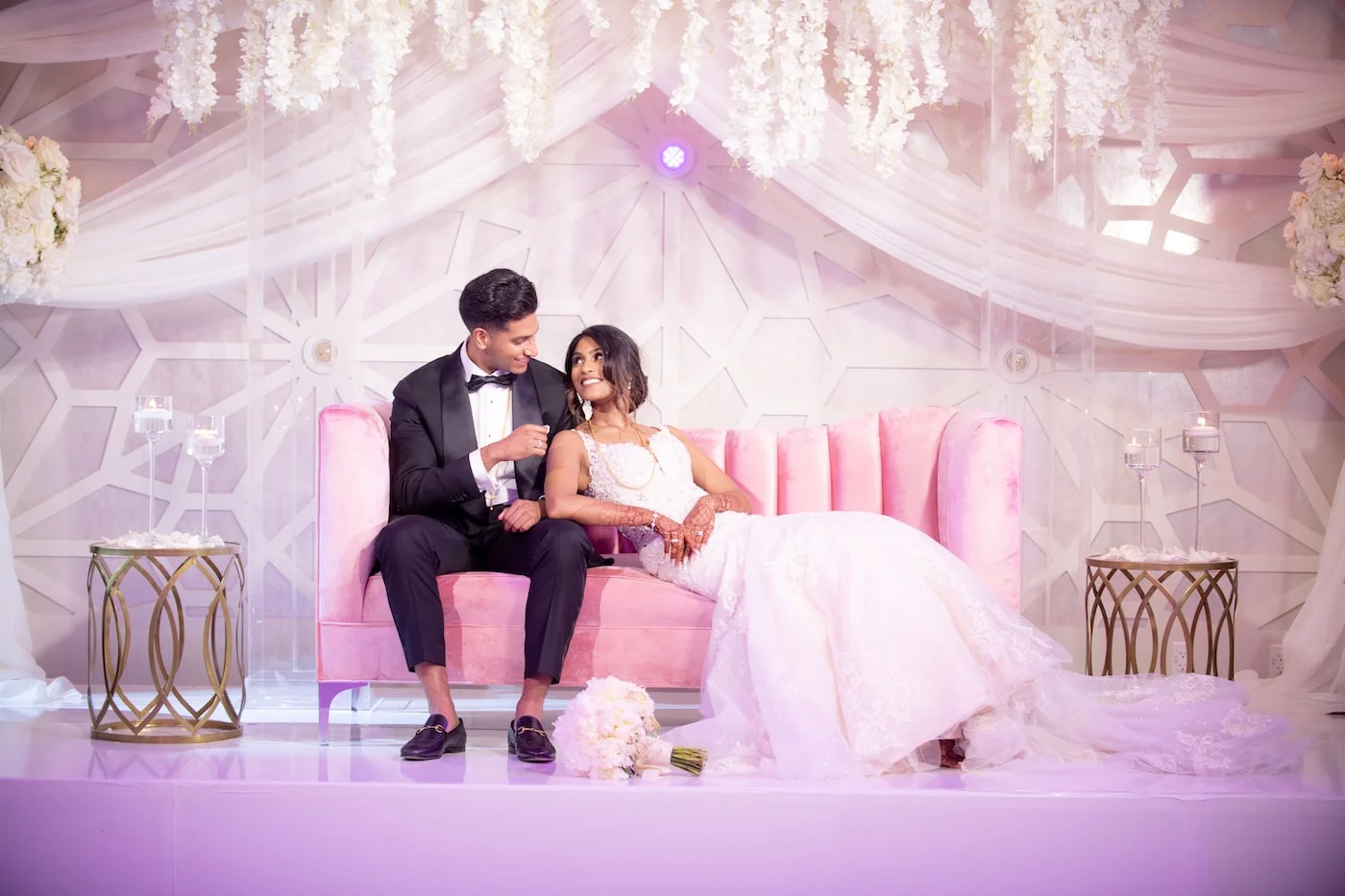 Bride and groom seated together on a pink loveseat on a draped reception stage