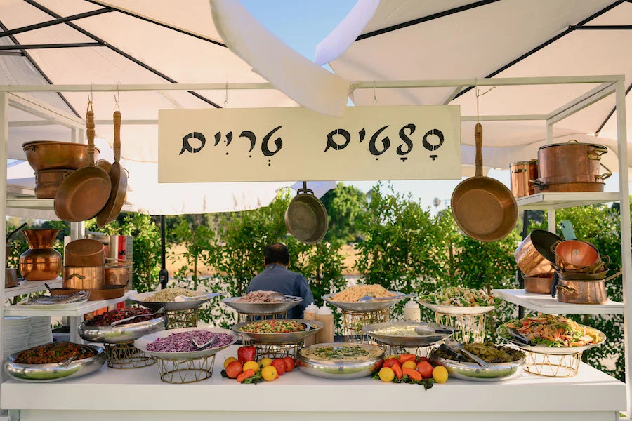 Outdoor catering station with copper cookware and a Hebrew menu sign over food trays