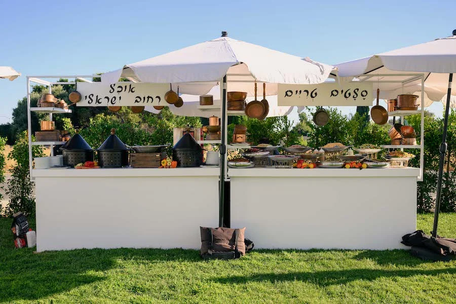Outdoor buffet stations under white umbrellas with copper pots and Hebrew signage