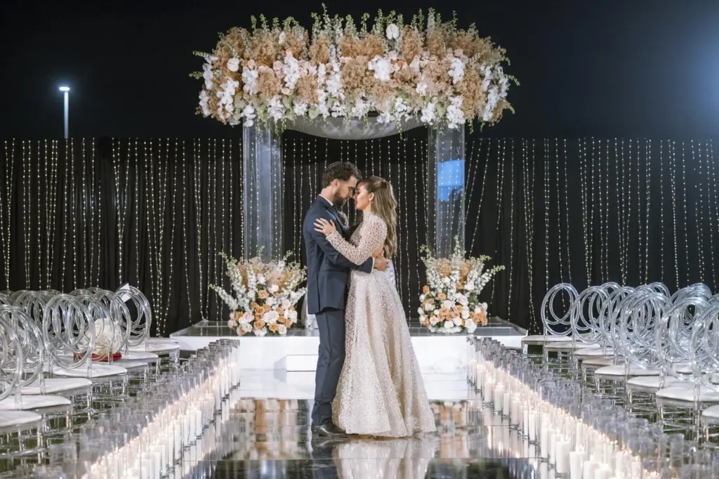 Bride and groom embracing under a floral chuppah on a candlelit aisle at night