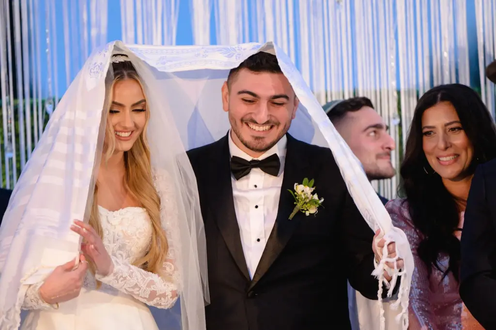 Bride and groom smiling together under a tallit during a Jewish wedding ceremony