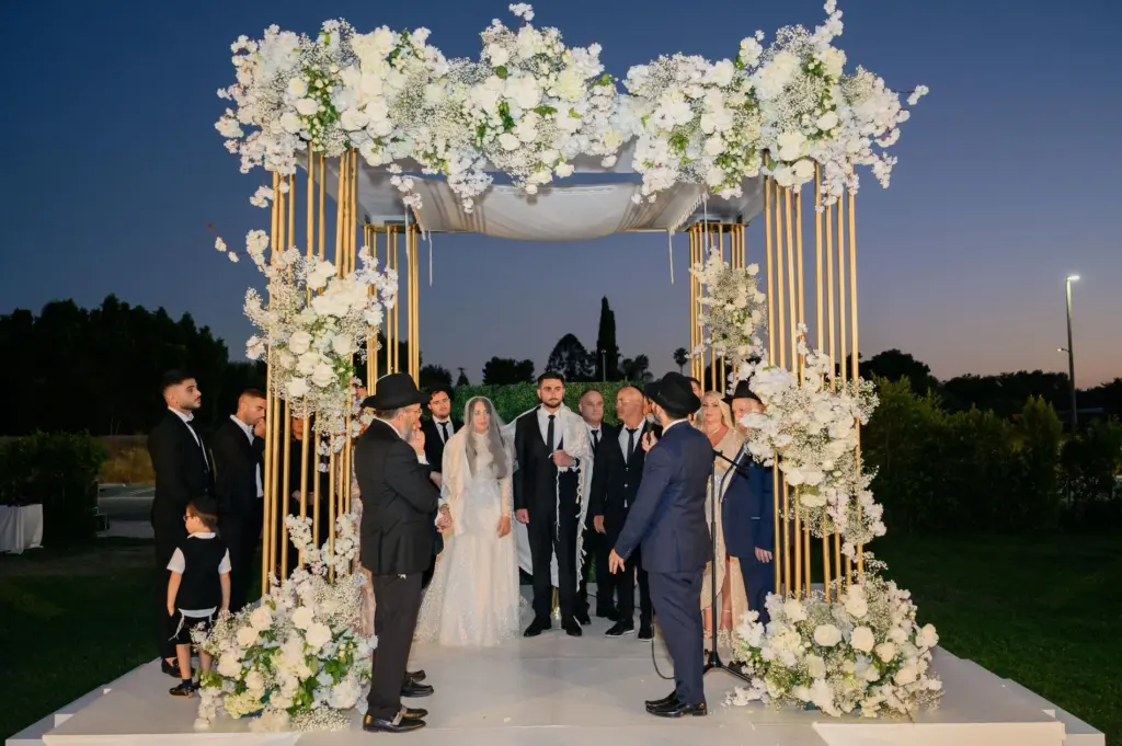 Guests in formal attire gathered around a gold-framed floral chuppah at an evening ceremony