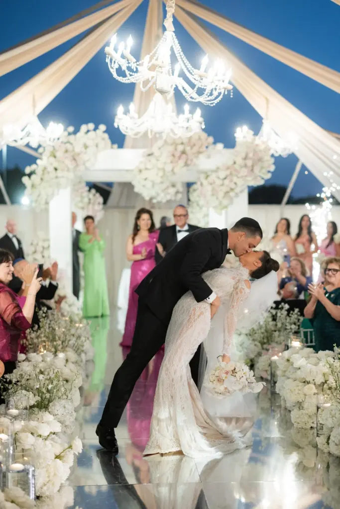 Bride and groom share a dip kiss under a floral arch as guests applaud