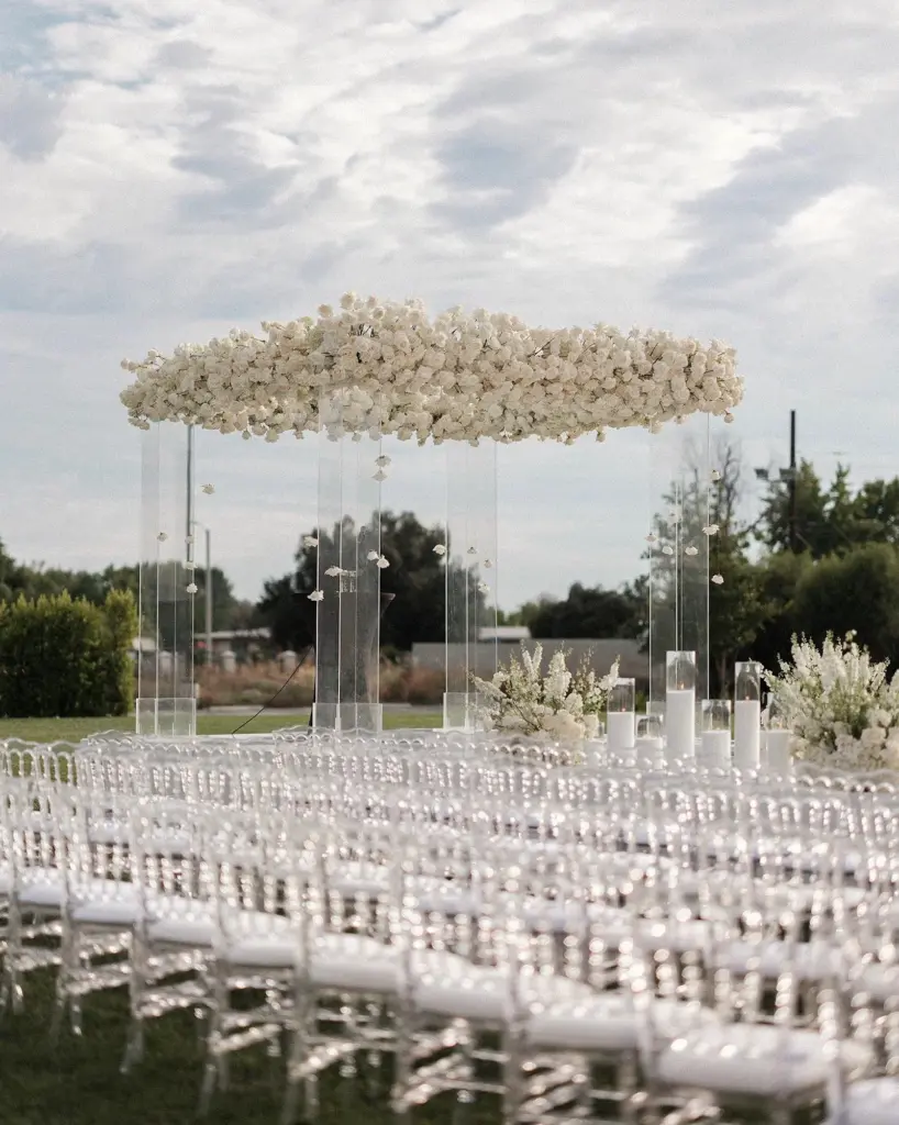 Floral canopy on clear acrylic columns above rows of clear chiavari chairs outdoors