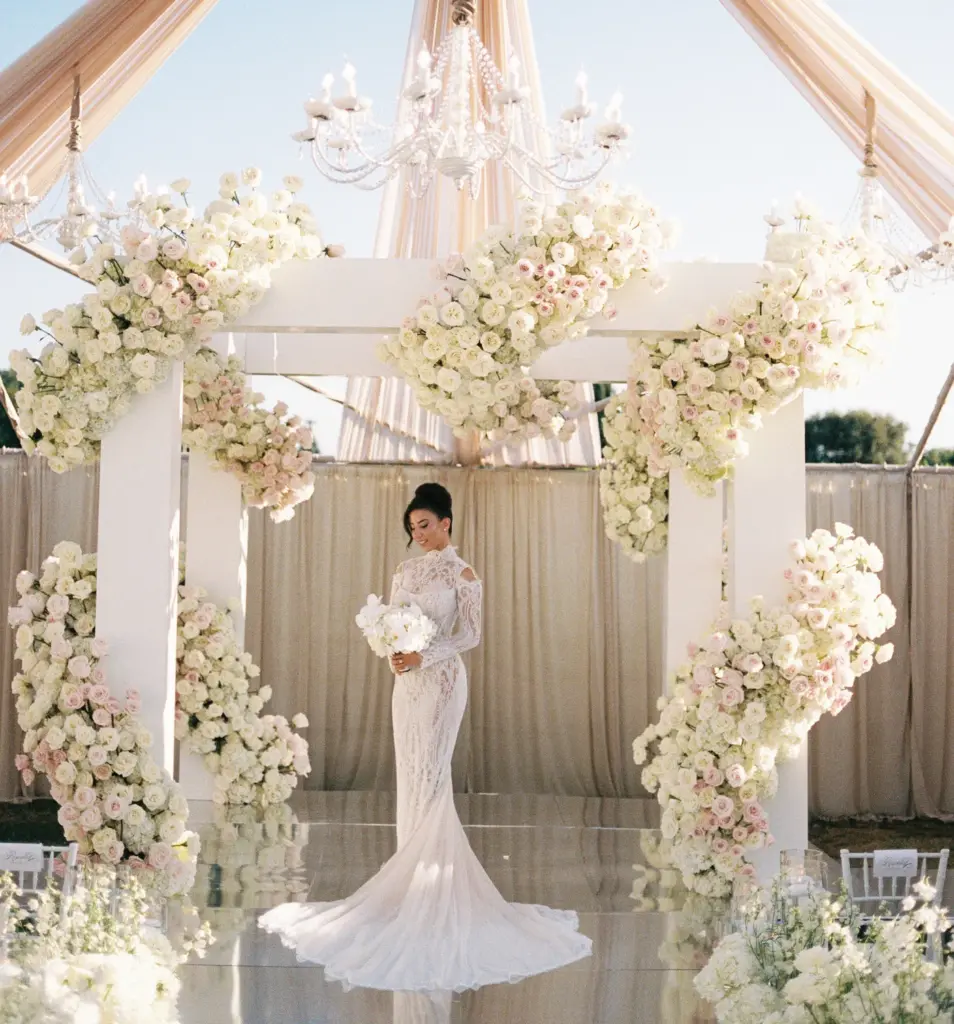 Bride in a lace gown standing under a floral arch and crystal chandelier
