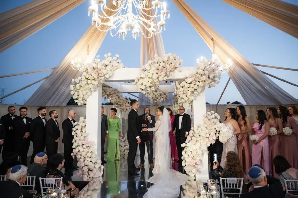 Wedding party and guests under a crystal chandelier and floral chuppah at an evening ceremony