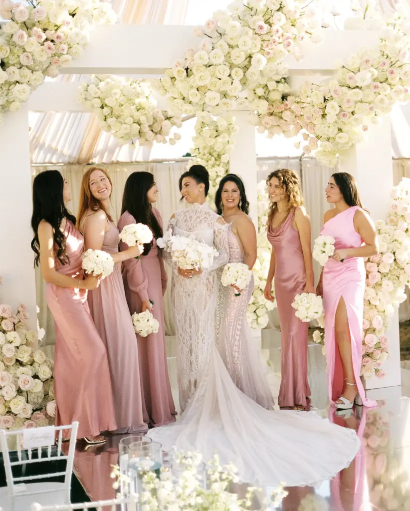 Bride and bridesmaids in pink gowns laughing together under a floral arch