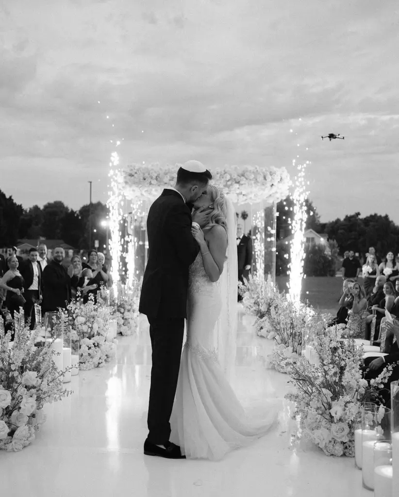 Black-and-white photo of bride and groom kissing under a floral chuppah with sparklers and a drone overhead