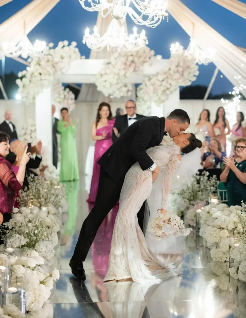Bride and groom share a dip kiss under a tented floral canopy as guests applaud